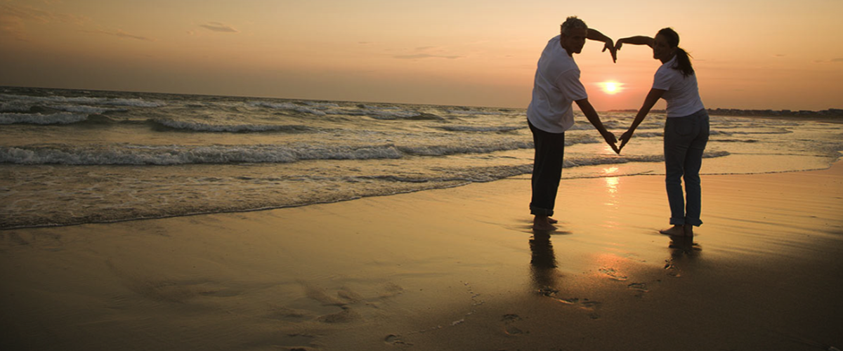 Image of a couple make a heart shape with their arms against the sunset on a beach.